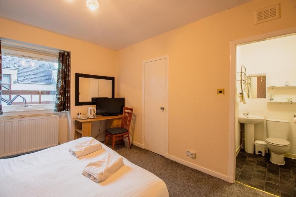 Habitación de hotel con cama, escritorio con computadora, silla, televisor y baño con inodoro y lavamanos.