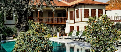 A poolside area with a clear view of a building with a tiled roof and balconies, surrounded by lush greenery and orange trees.