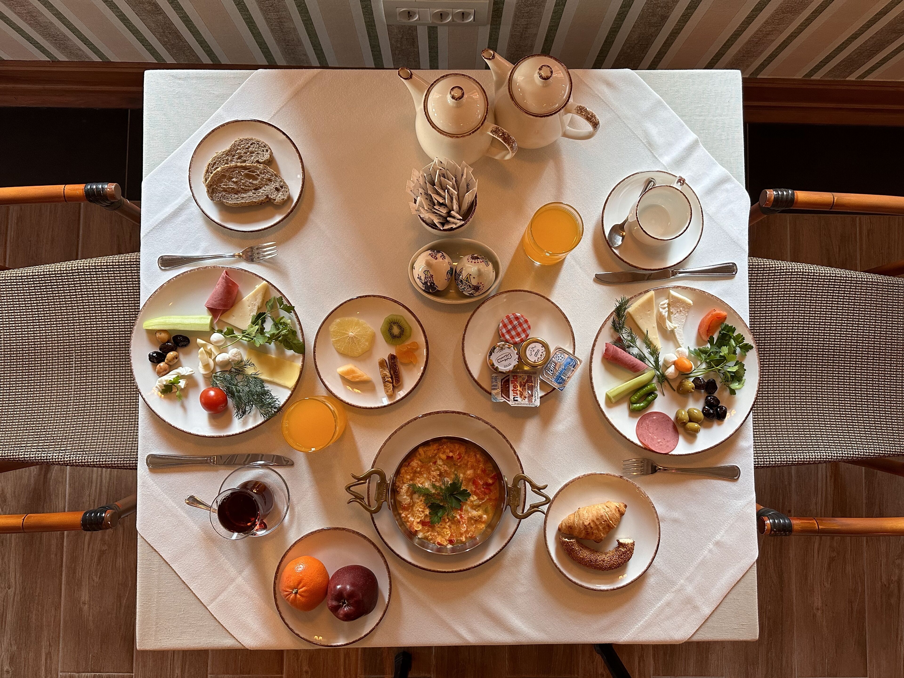 Una escena de desayuno con diversos platos, una tetera y un vaso de jugo de naranja sobre una mesa.