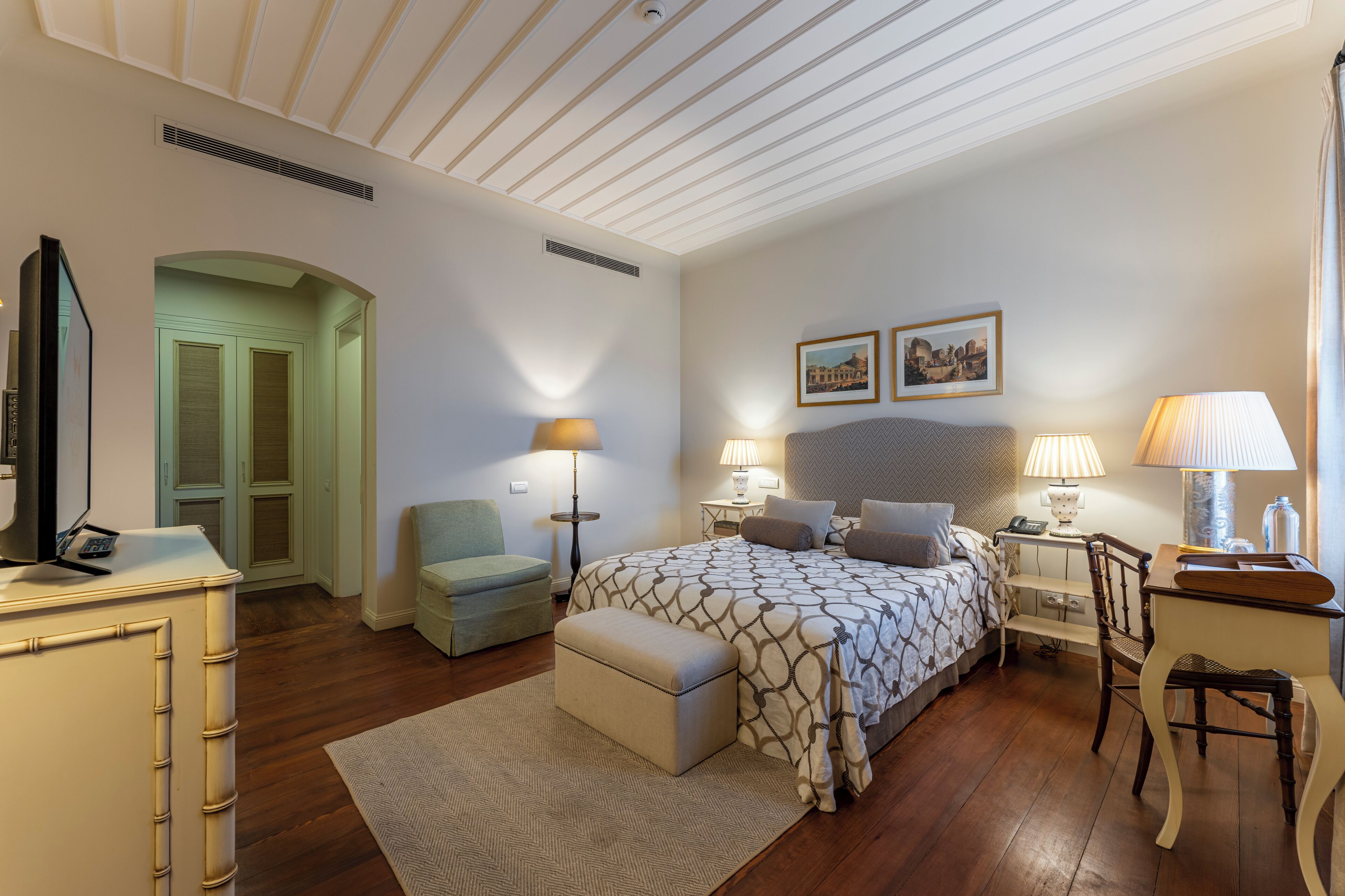 A bedroom with a large bed, bedside tables, a desk, a chair, a television, and a door leading to another room.