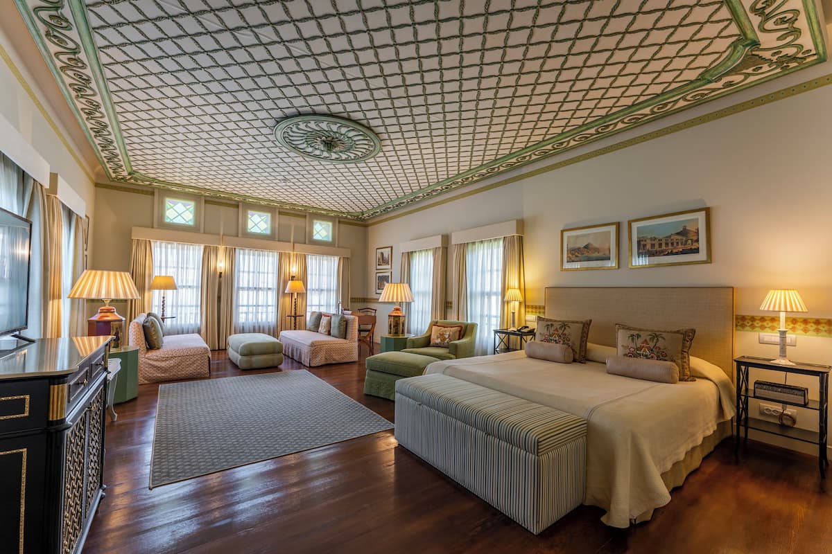 Une chambre d’hôtel spacieuse, dotée d’un grand lit, de deux fauteuils, d’un canapé, d’un bureau et d’une télévision. La chambre a un plafond haut orné d’un motif décoratif et comprend plusieurs fenêtres.