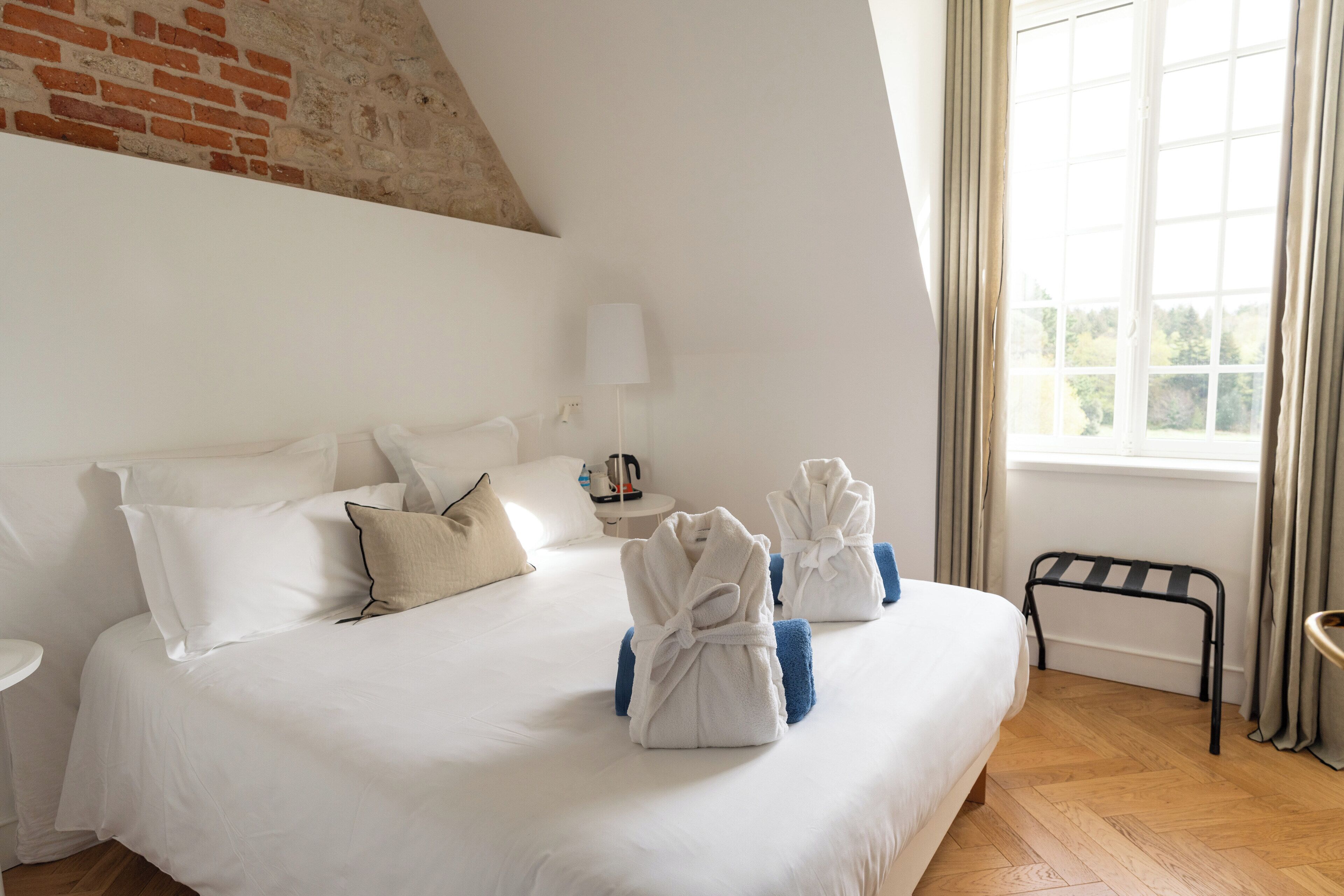Habitación de hotel con una cama grande, ropa de cama blanca, pared de ladrillo, ventana con cortinas y una mesita con una lámpara.