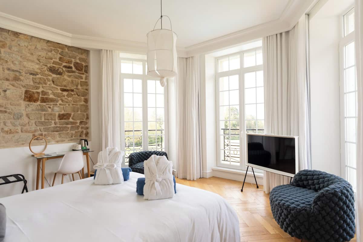 A hotel room with a large bed, a desk with a chair, a TV on a stand, and a stone wall.
