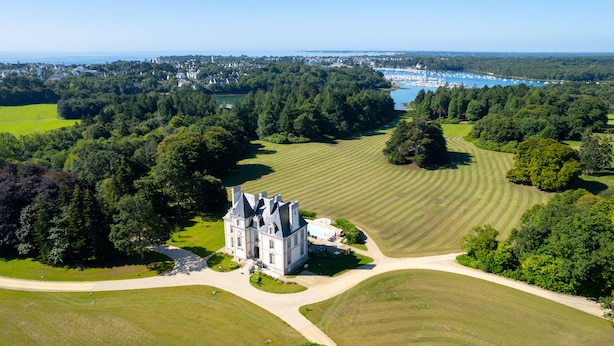 An aerial view of a large estate with a prominent castle-like building surrounded by expansive green fields and a marina with boats.