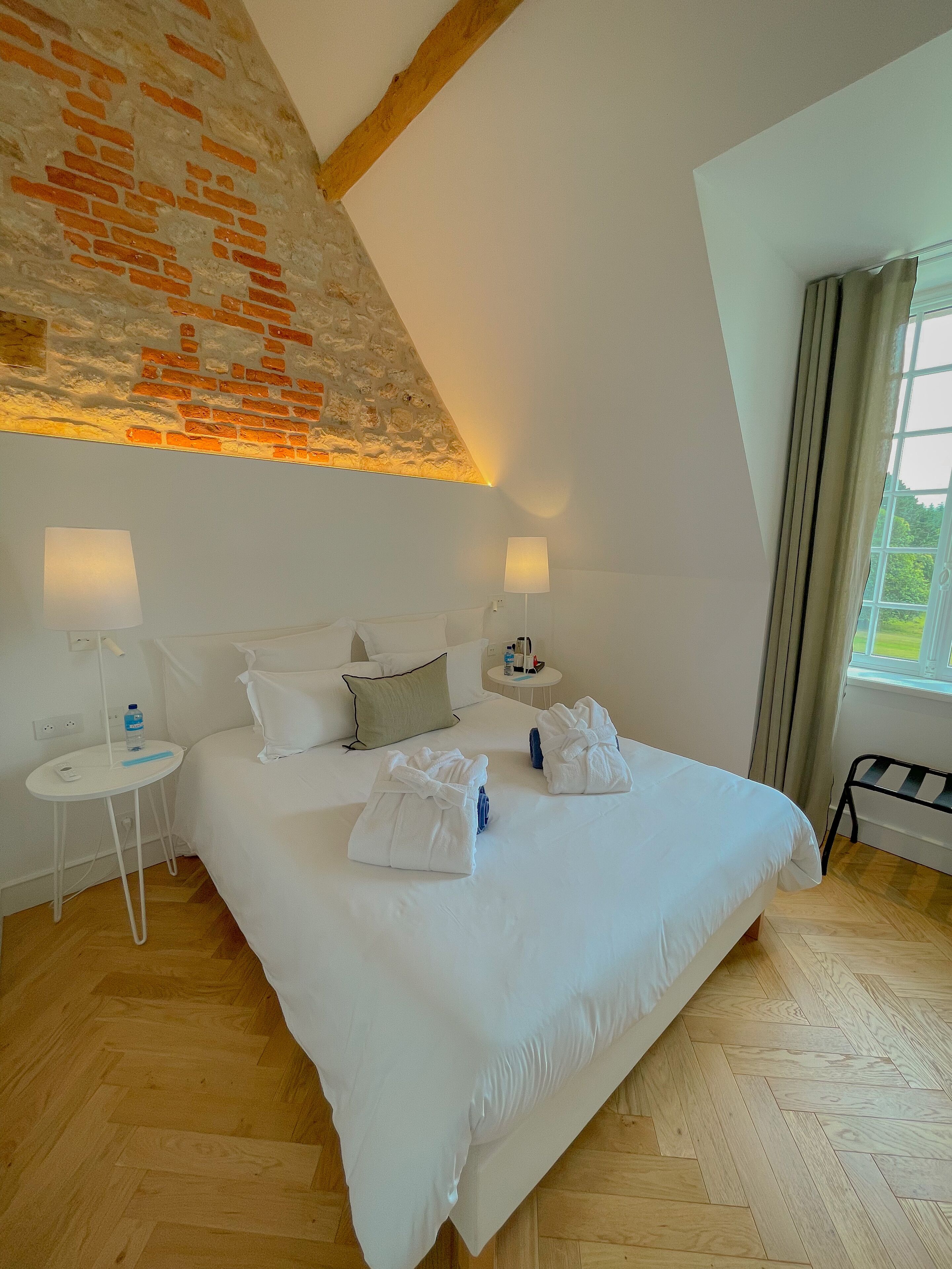 Ein Schlafzimmer mit einem großen Bett, zwei Nachttischen, einer Lampe und Blick auf Grünflächen.