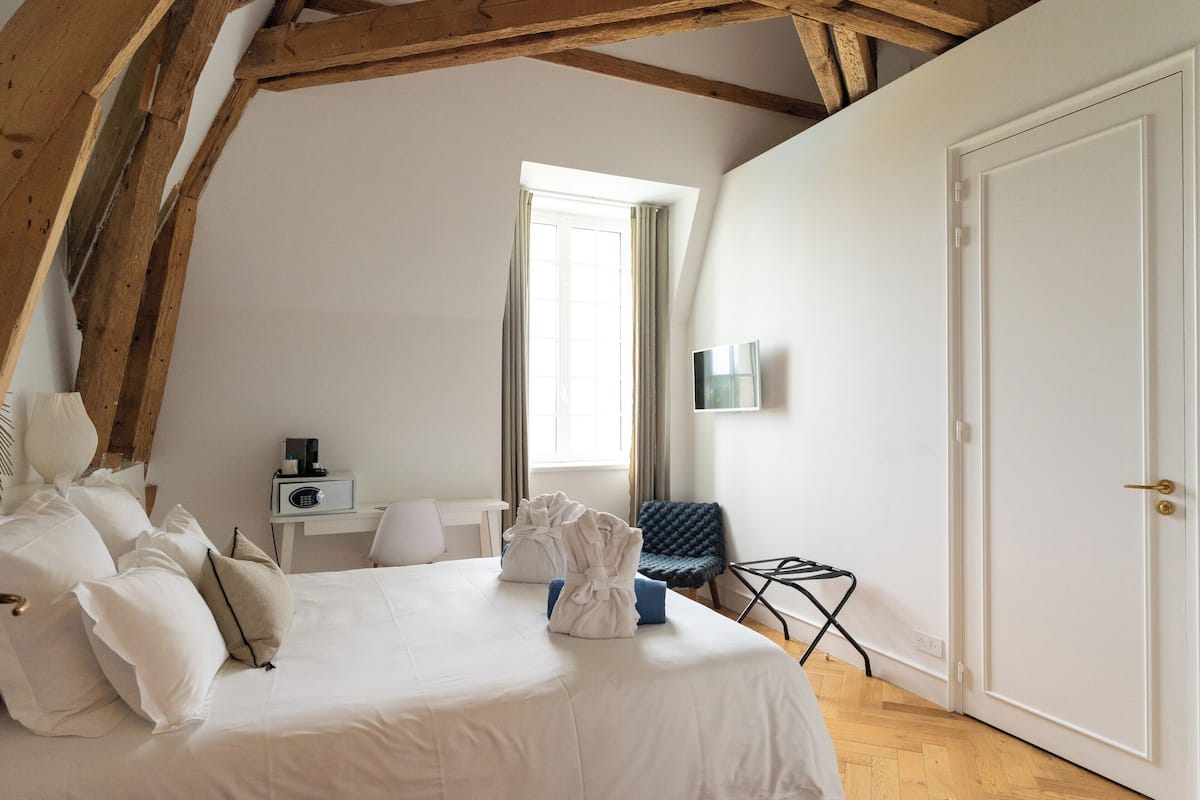 A bedroom with a large bed, a bedside table with a radio, a chair, a window with curtains, and a door.