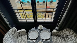 A dining area with a view of a swimming pool, two patterned chairs, a table set with plates, cutlery, and a basket of bread and pastries.