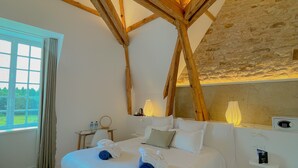A bedroom with a wooden ceiling structure, a bed with white linens, a bedside table with a lamp, and a view of greenery outside.