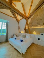 Une chambre avec une structure de plafond en bois, un lit avec du linge de lit blanc, une table de chevet avec une lampe, et une vue sur la verdure à l’extérieur.