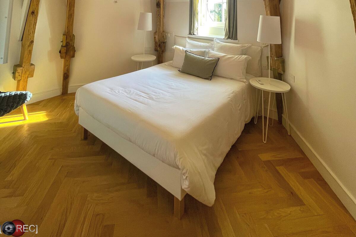 A bedroom with a sloped ceiling, wooden beams, a bed with white linens, two bedside tables with lamps, and a window with curtains.