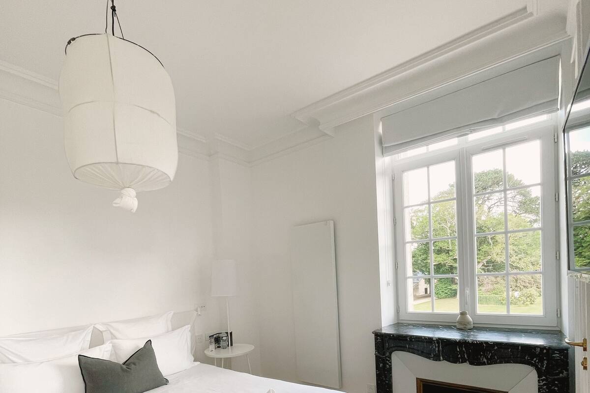 A bedroom with a large bed, a window with a view of greenery, a fireplace, and a hanging light fixture.
