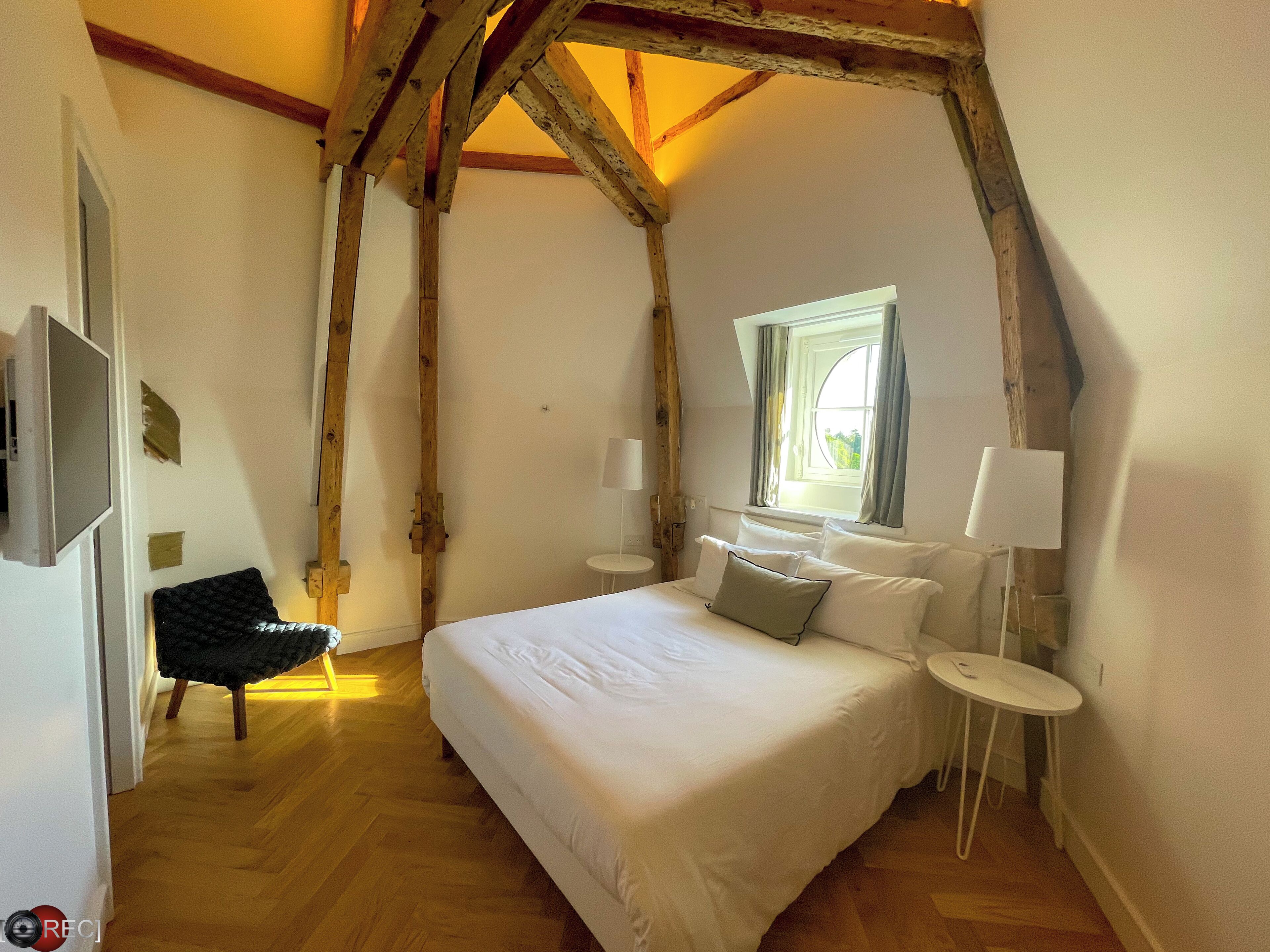 Un dormitorio con una cama grande, dos mesitas de noche, una ventana con cortinas, vigas de madera y una silla.