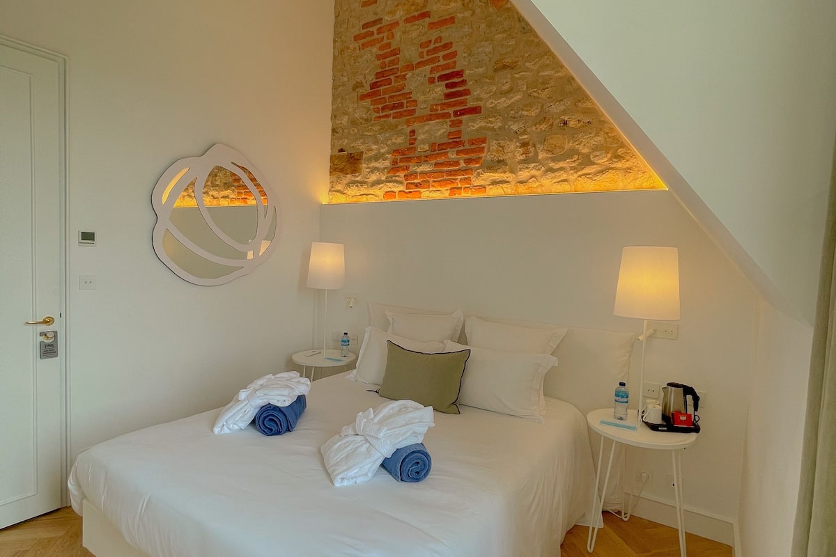 A hotel room with a large bed, a wooden ceiling, a stone wall, a round mirror, two bedside tables with lamps, and a small table with a kettle and a bottle.