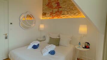Une chambre d’hôtel avec un grand lit, un plafond en bois, un mur en pierre, un miroir rond, deux tables de chevet avec des lampes et une petite table avec une bouilloire et une bouteille.