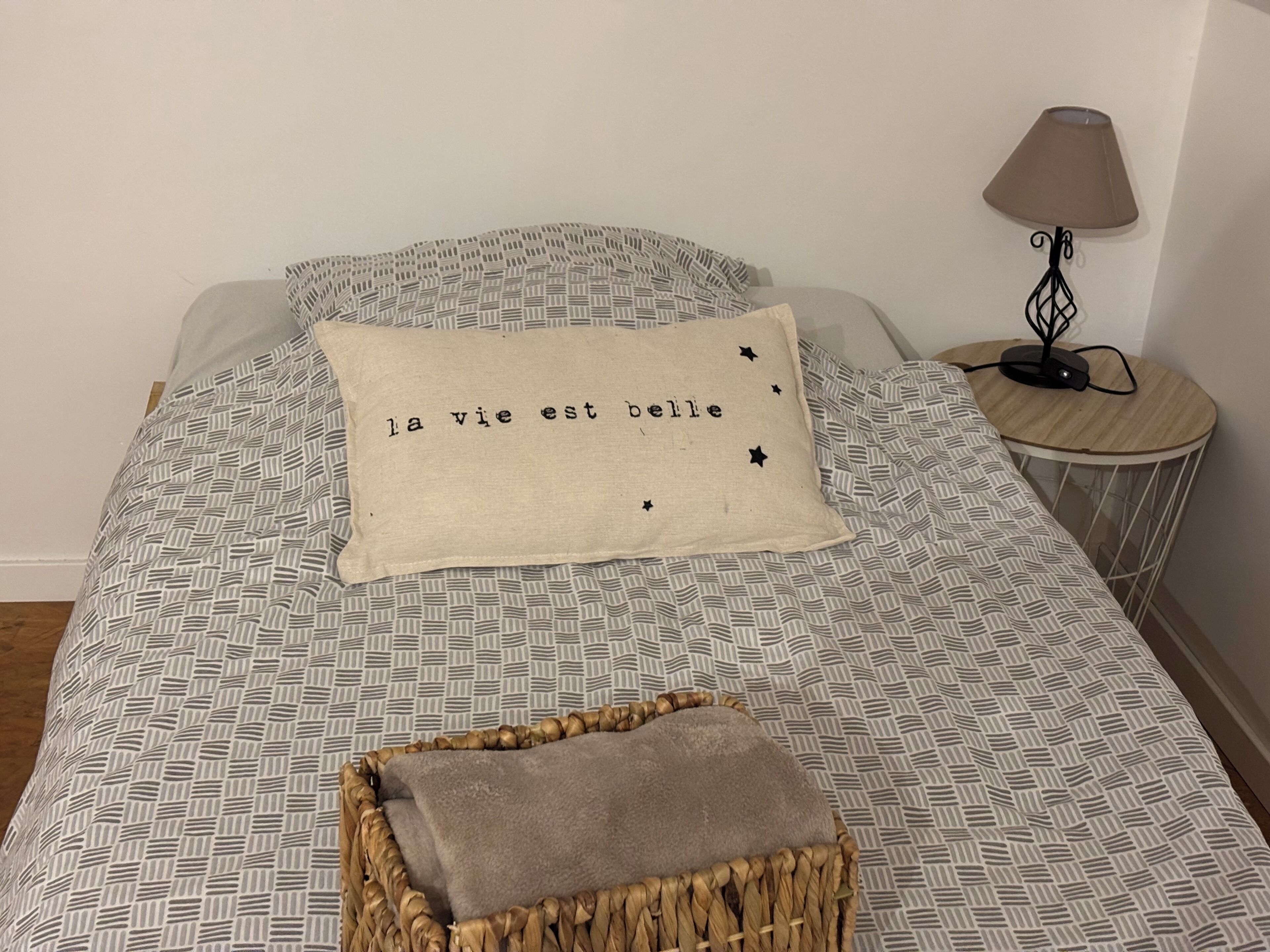 Una cama bien hecha con una colcha estampada, un almohadón con el texto "la vie est belle", una pequeña cesta tejida y una lámpara de noche sobre una mesita de madera.