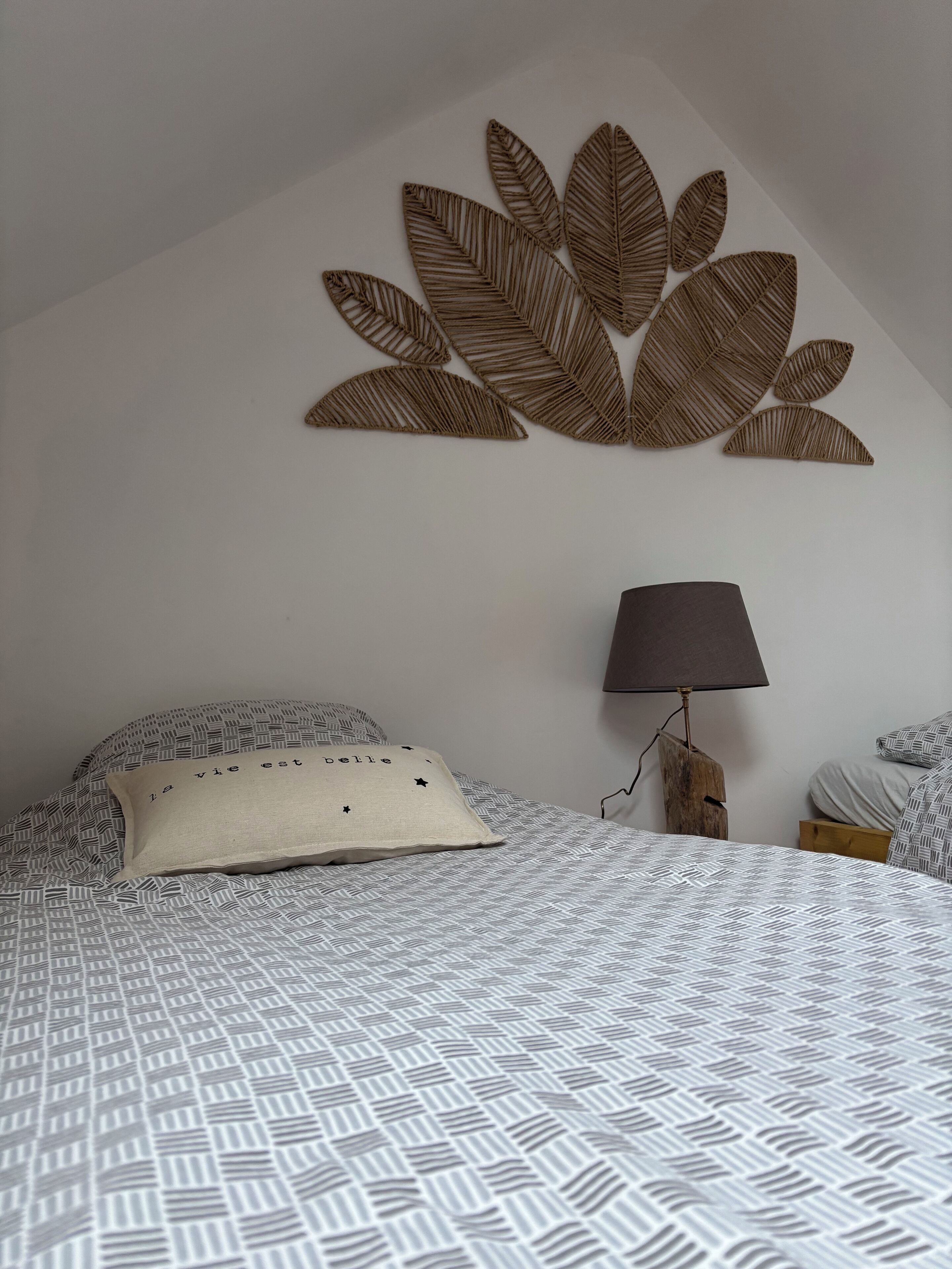 Una camera da letto con una coperta a motivi, un cuscino con scritte, una decorazione a forma di foglia appesa al muro, una lampada e un comodino.