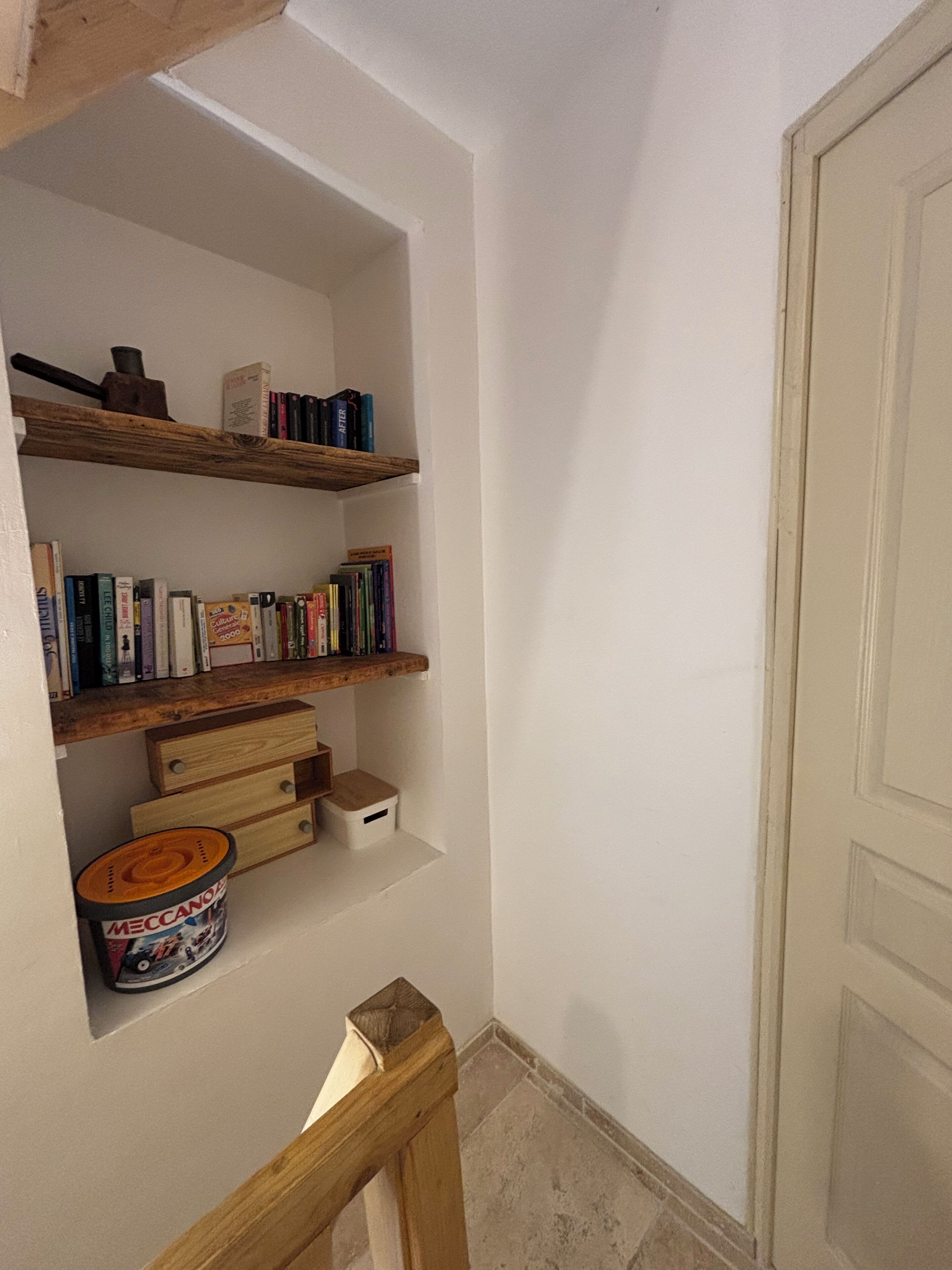 Um corredor estreito com paredes brancas e uma porta de madeira. Há duas estantes de madeira com livros e um pequeno armário de madeira.