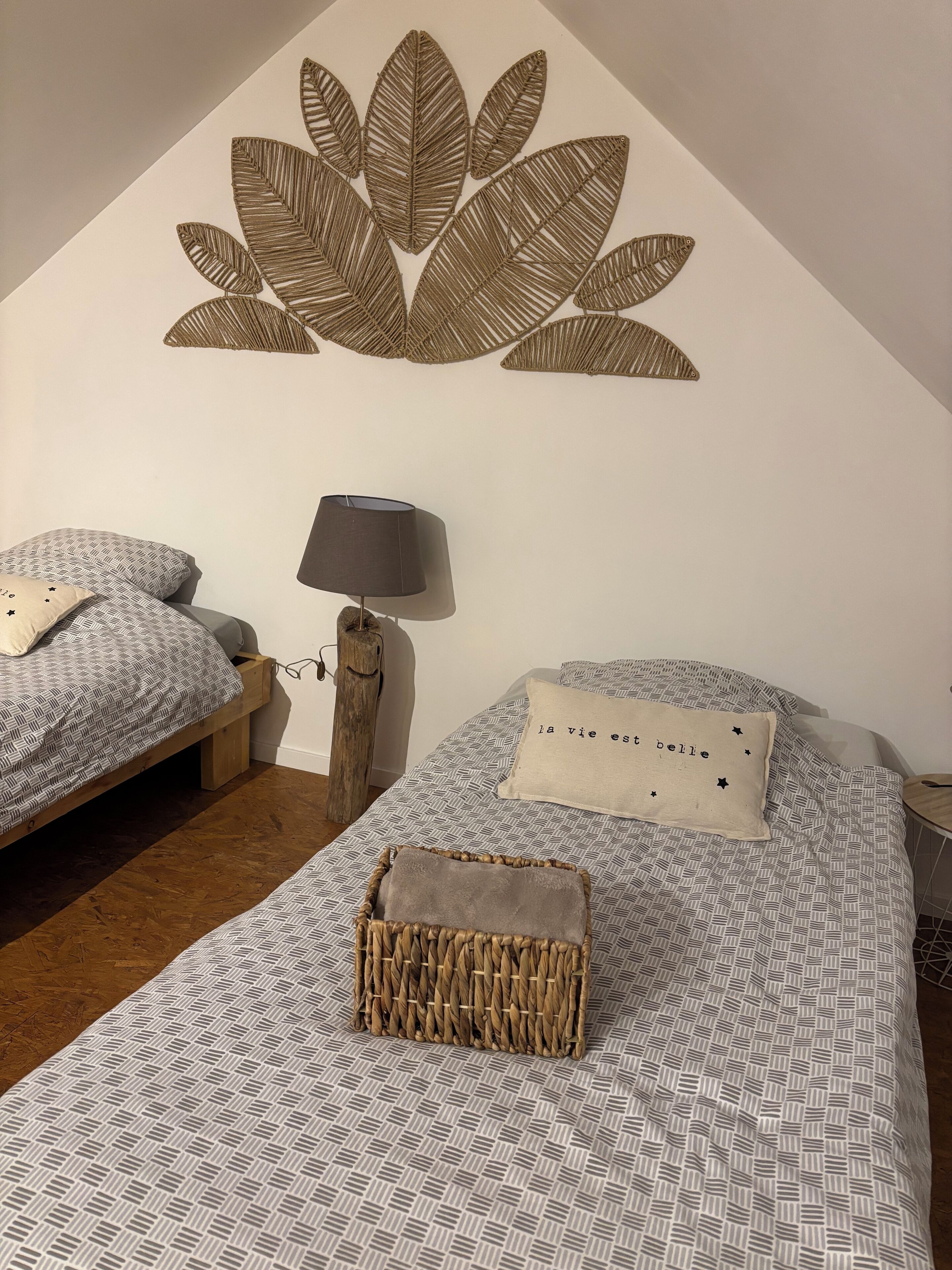Une chambre avec deux lits, une décoration murale en forme de feuille, une lampe et un panier en osier posé sur l’un des lits.