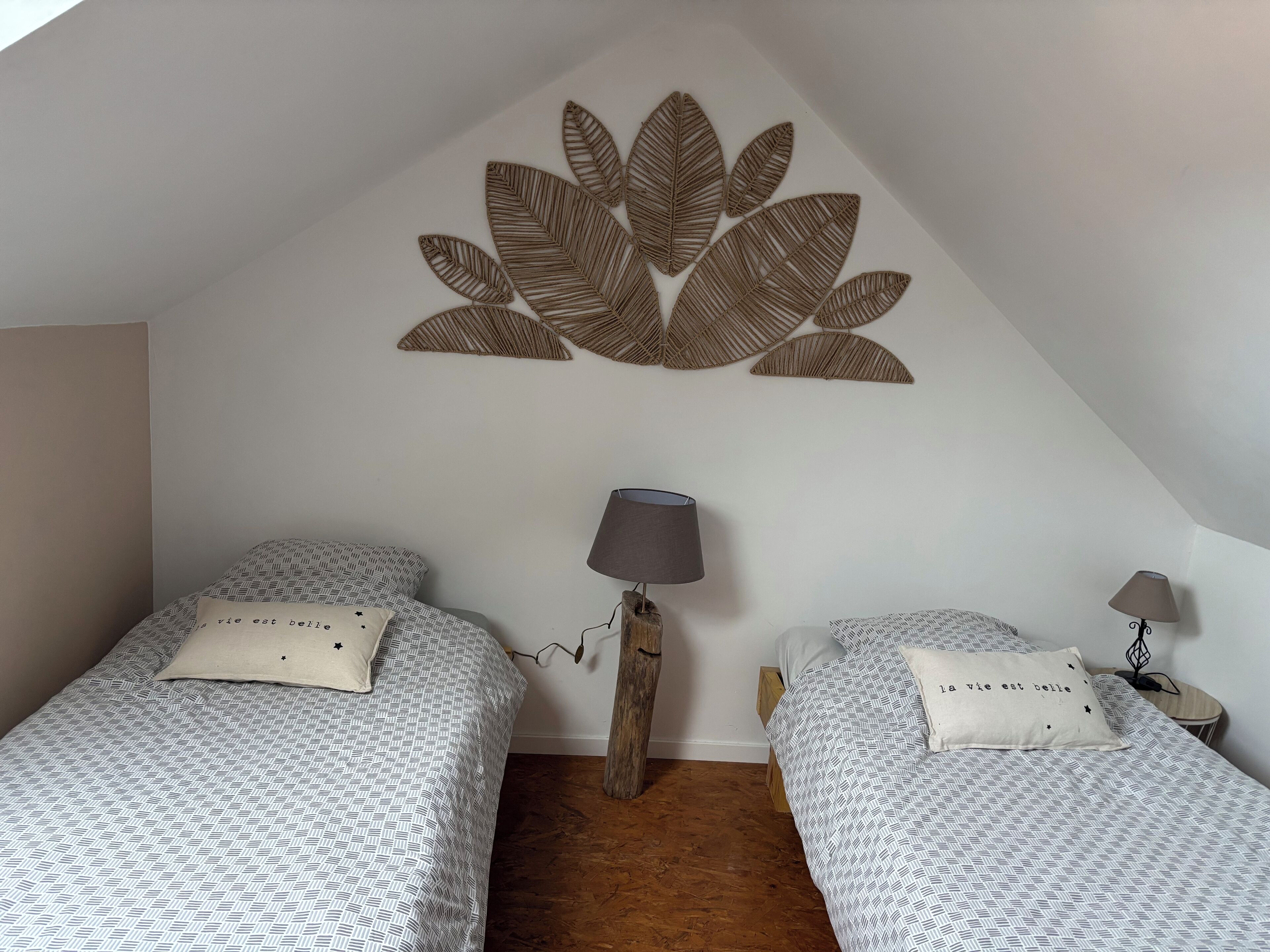 Ein Schlafzimmer mit zwei Betten, jeweils mit gemusterten Bettbezügen und einem Kissen mit Text. Ein Nachttisch aus Holz mit einer Lampe befindet sich zwischen den Betten.