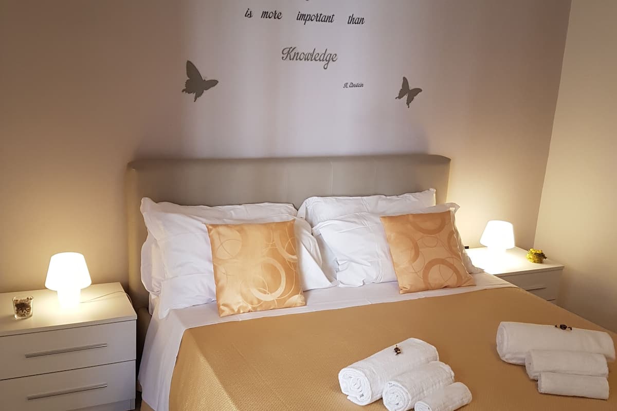A neatly made bed with white and gold bedding, two nightstands with lamps, and a wall with a quote about imagination and knowledge.