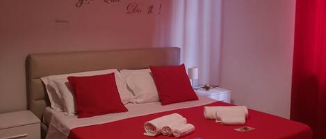 A neatly made bed with red and white bedding, a red throw blanket, and rolled towels. There is a wall with a motivational quote.