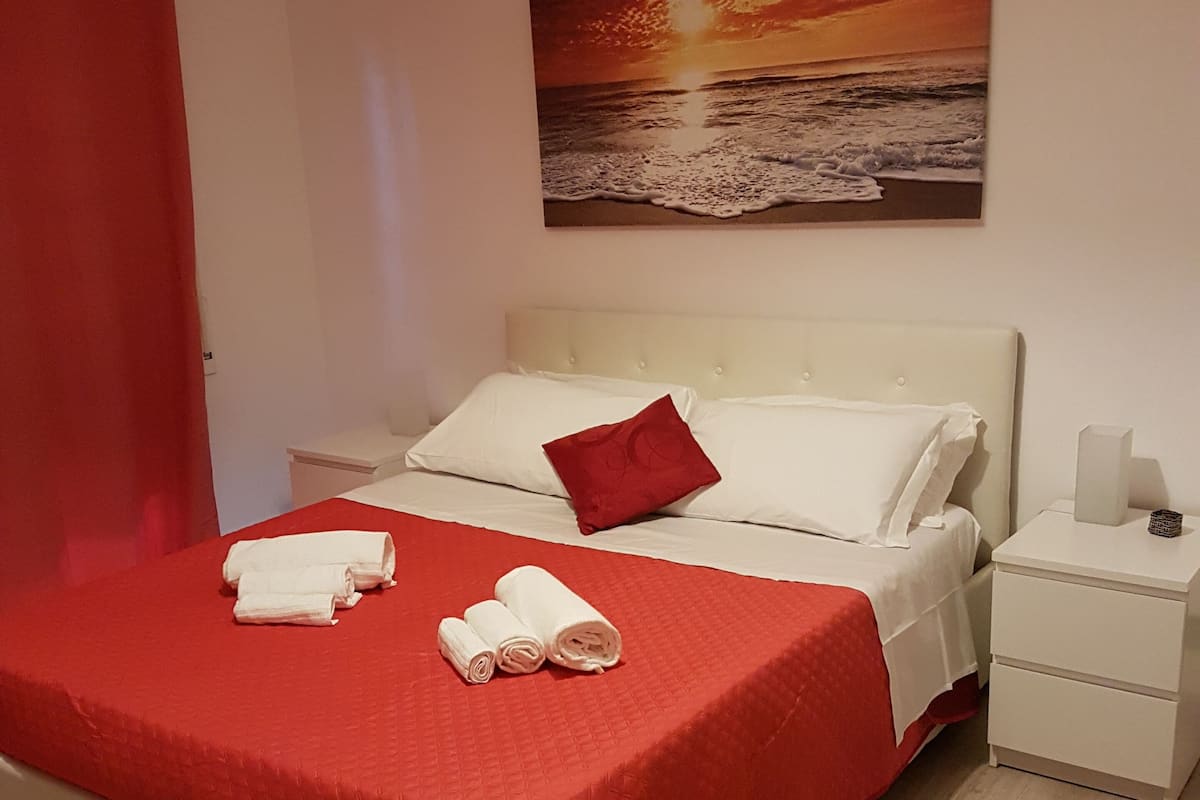 A neatly made bed with a red blanket and white pillows, a white nightstand with a lamp, and a painting of a sunset over the ocean on the wall.