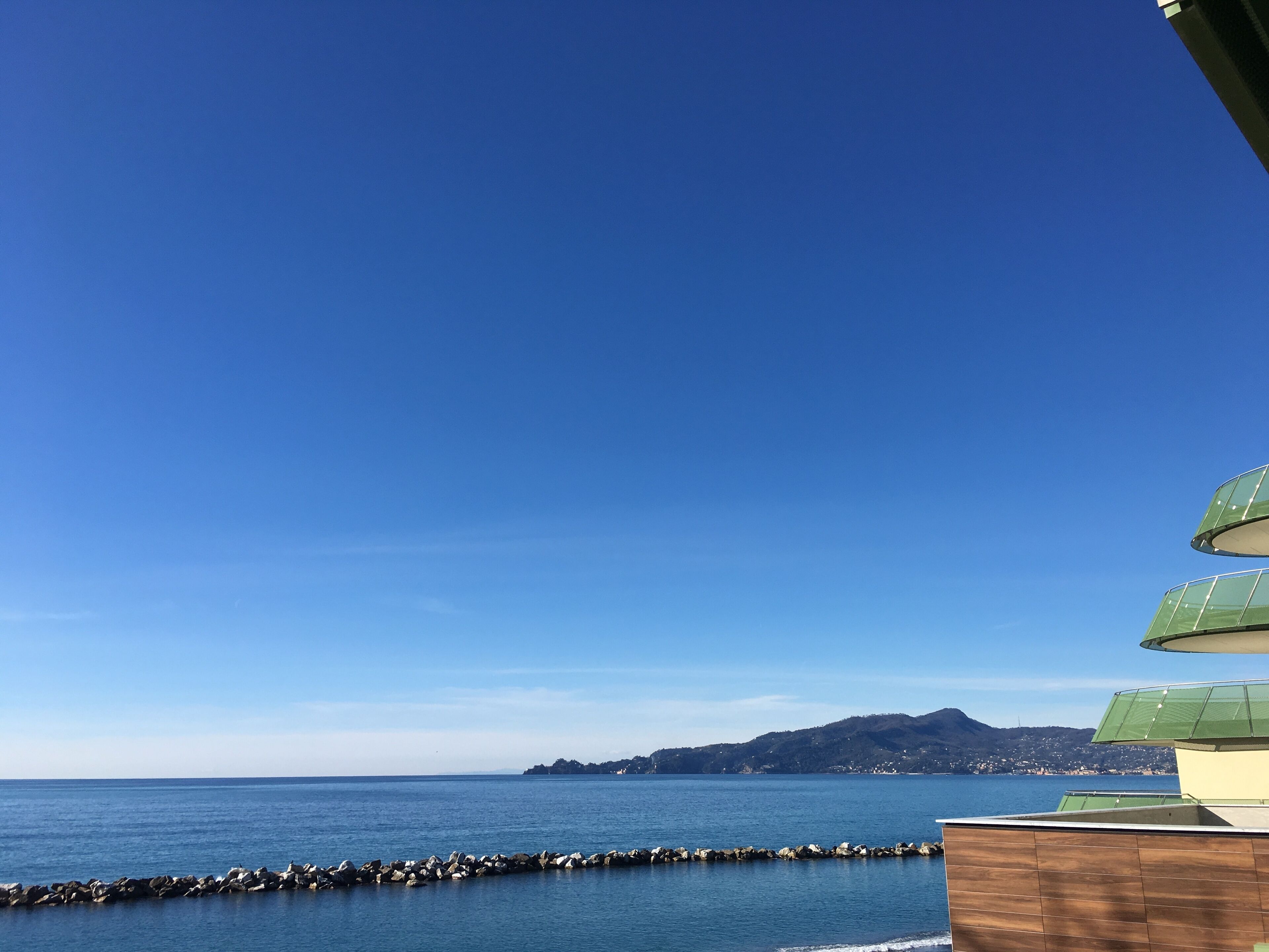 A coastal view with a clear sky, a rocky breakwater, and a distant mountain range.