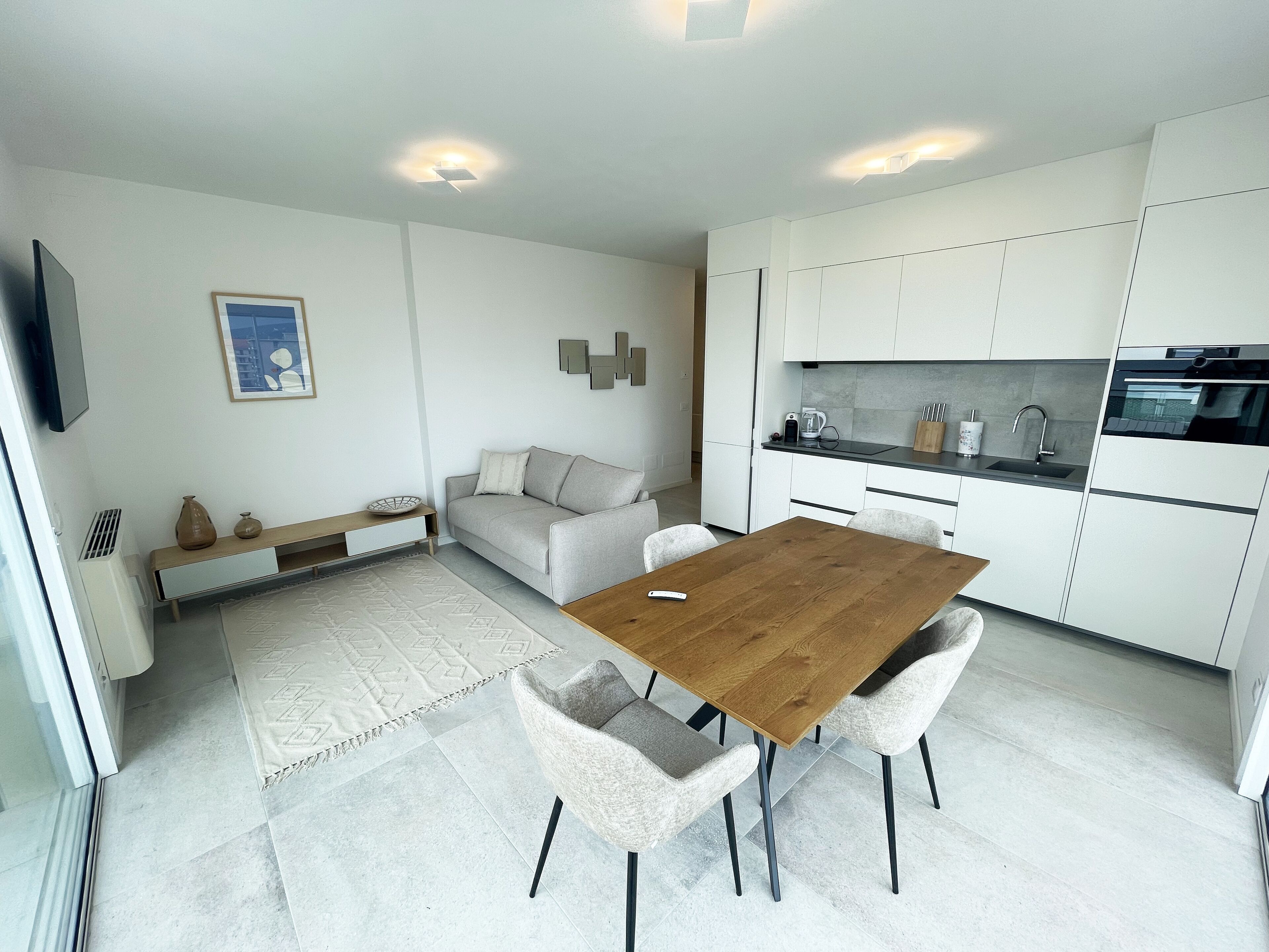 A modern kitchen with white cabinets, a dining area with a wooden table and chairs, a sofa, and a small rug.