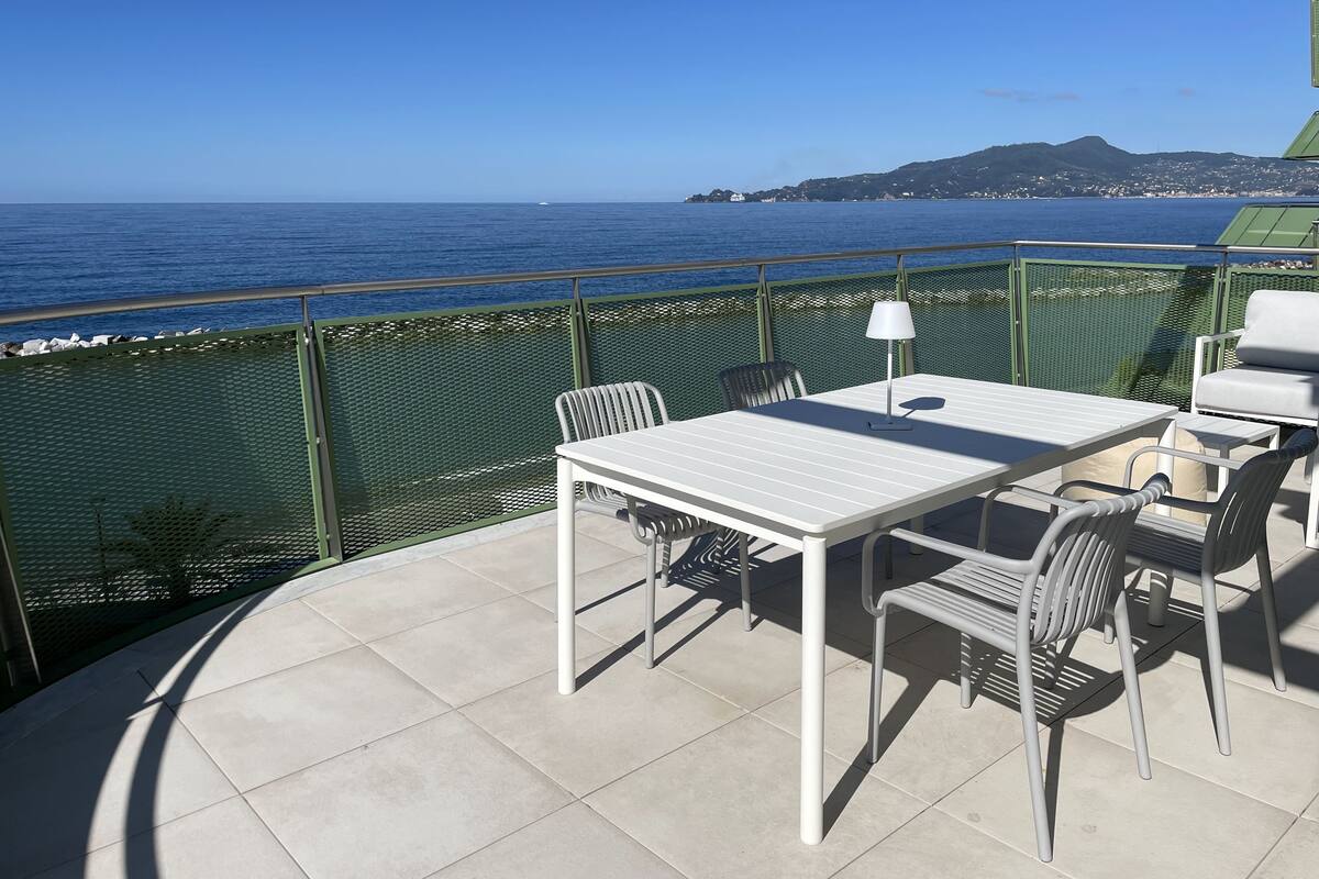 A balcony with a white table and chairs, a metal railing, and a view of the sea and distant hills.