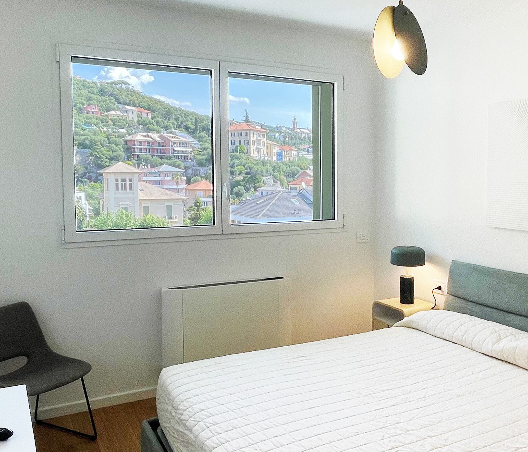 A bedroom with a bed, a chair, a nightstand, a lamp, and a window with a view of buildings and greenery.