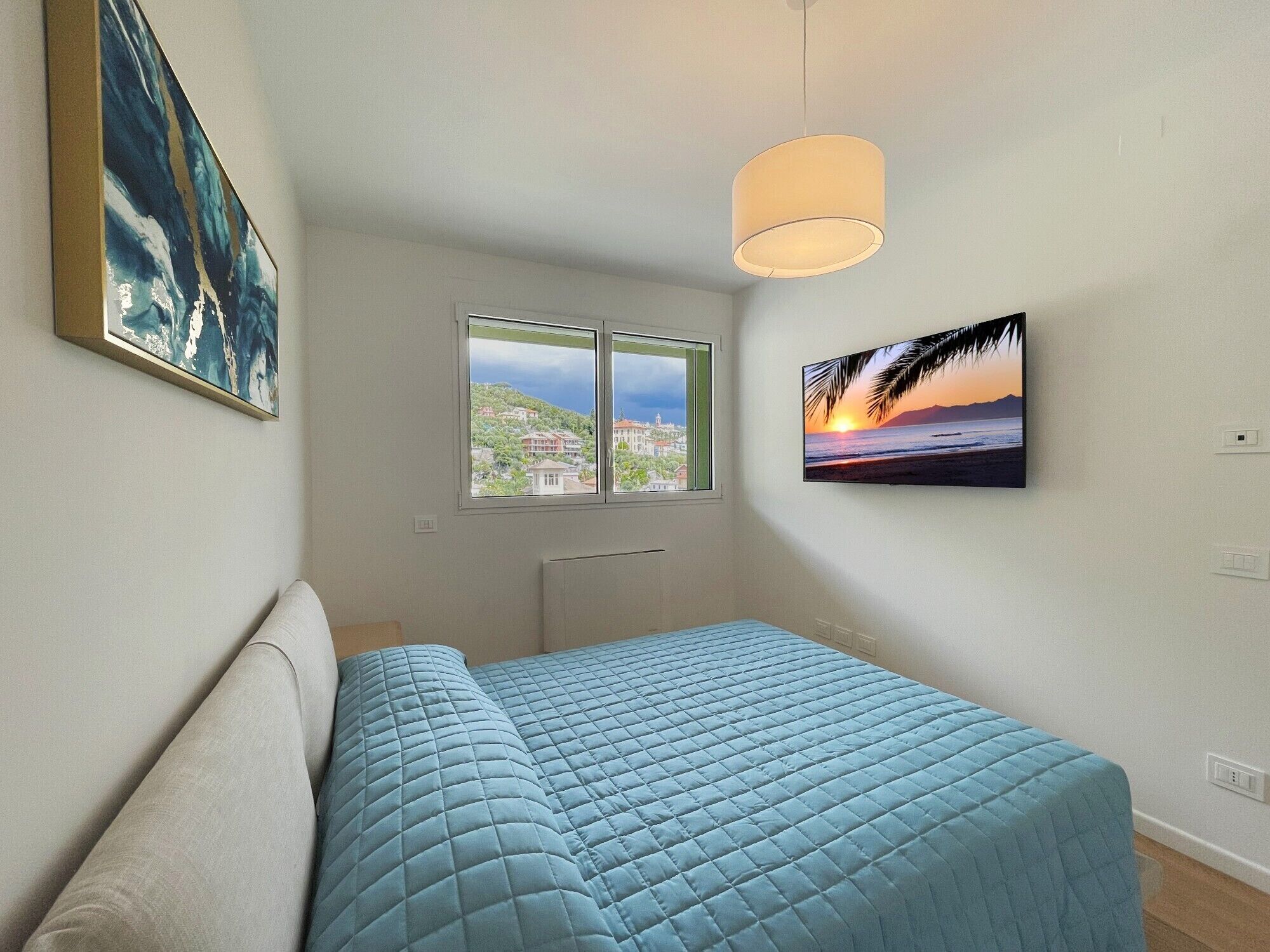 A bedroom with a bed, a window with a view of buildings, and a painting of a sunset.
