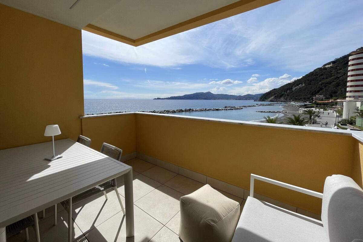 A balcony with a table and chairs, overlooking a coastal area with a clear sky and distant mountains.