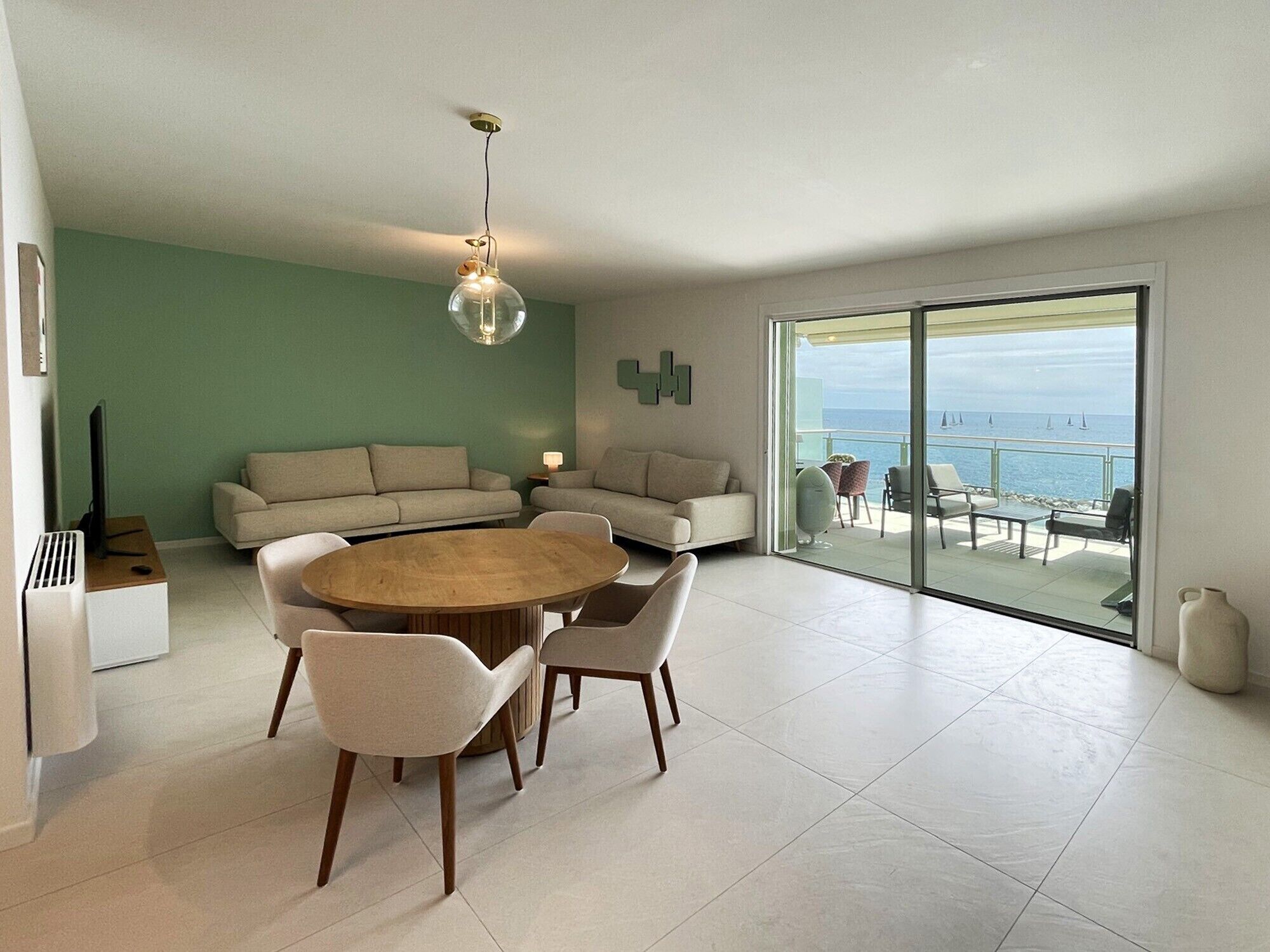 Ein modernes Wohnzimmer mit einem runden Holztisch und Stühlen, einer Couch und einer großen Schiebeglastür, die auf einen Balkon mit Meerblick führt.