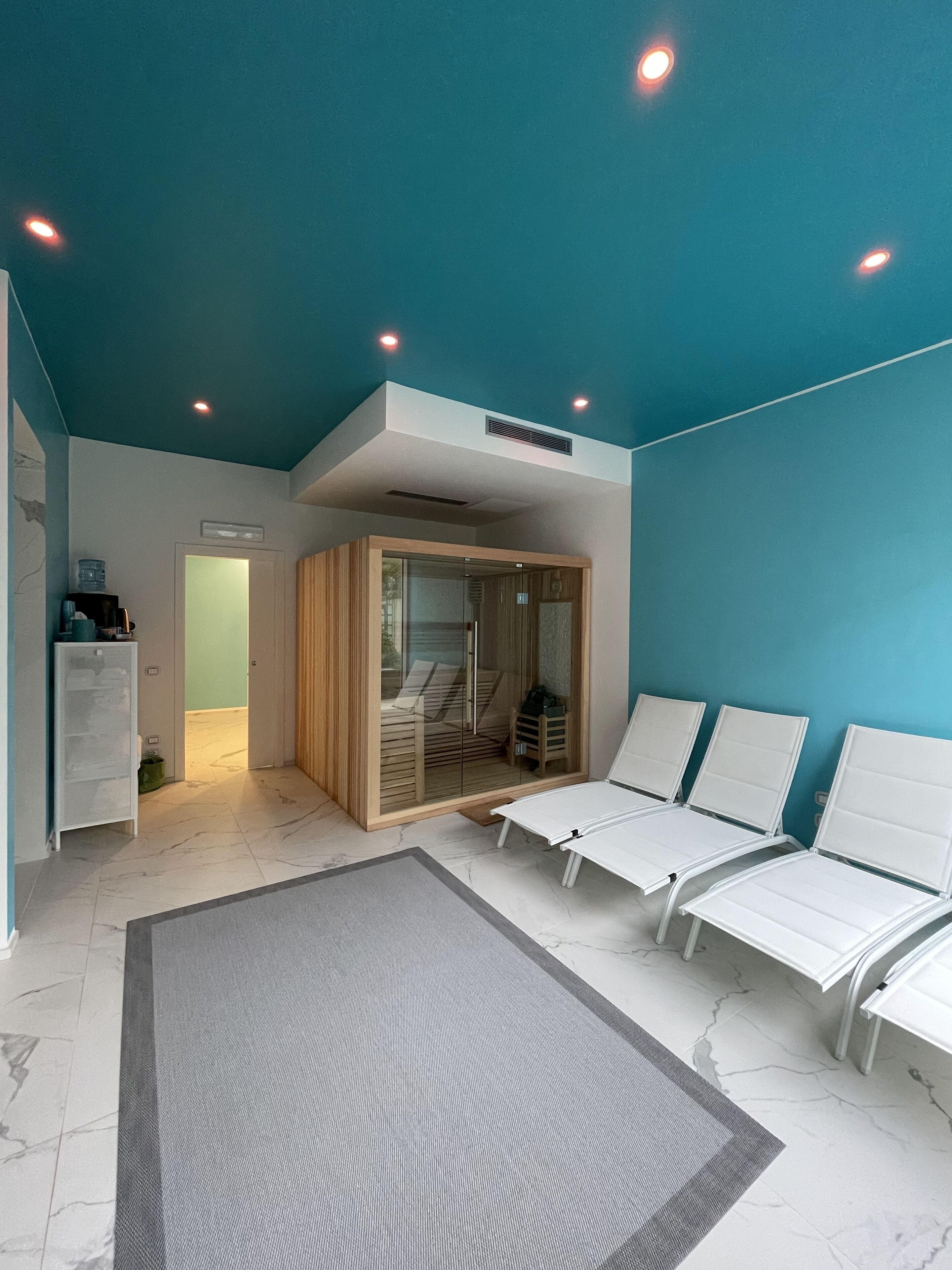 Un spa moderno con techo azul, sillones blancos, entrada por puerta de cristal y una pequeña cabaña de madera.