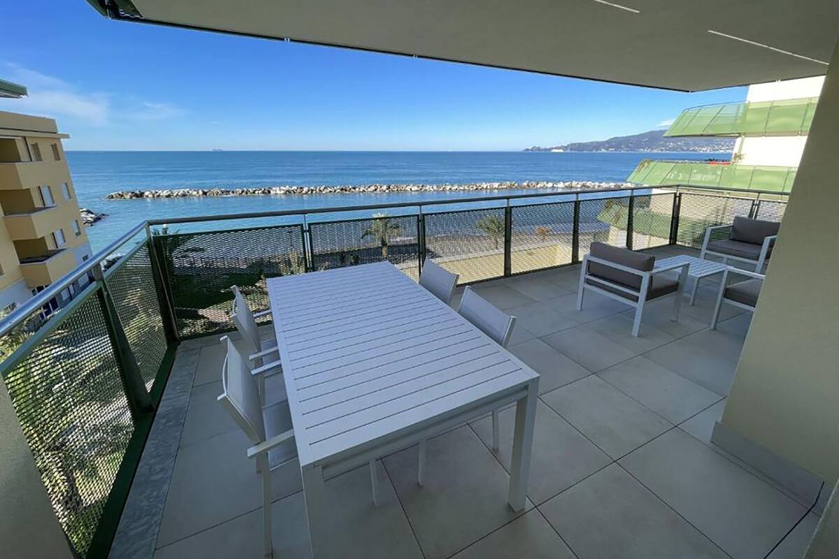 A balcony with a white table and chairs, overlooking a beach and the sea.