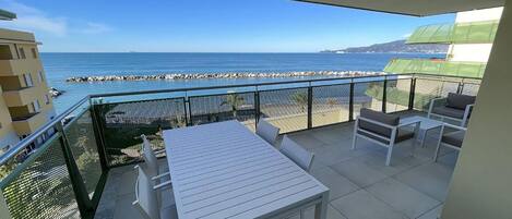 Ein Balkon mit weißem Tisch und Stühlen, der Blick auf einen Strand und das Meer freigibt.