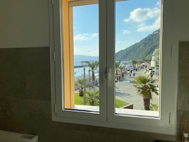 Una habitación con un ventanal grande que da a una playa con palmeras, un paseo marítimo y montañas al fondo.
