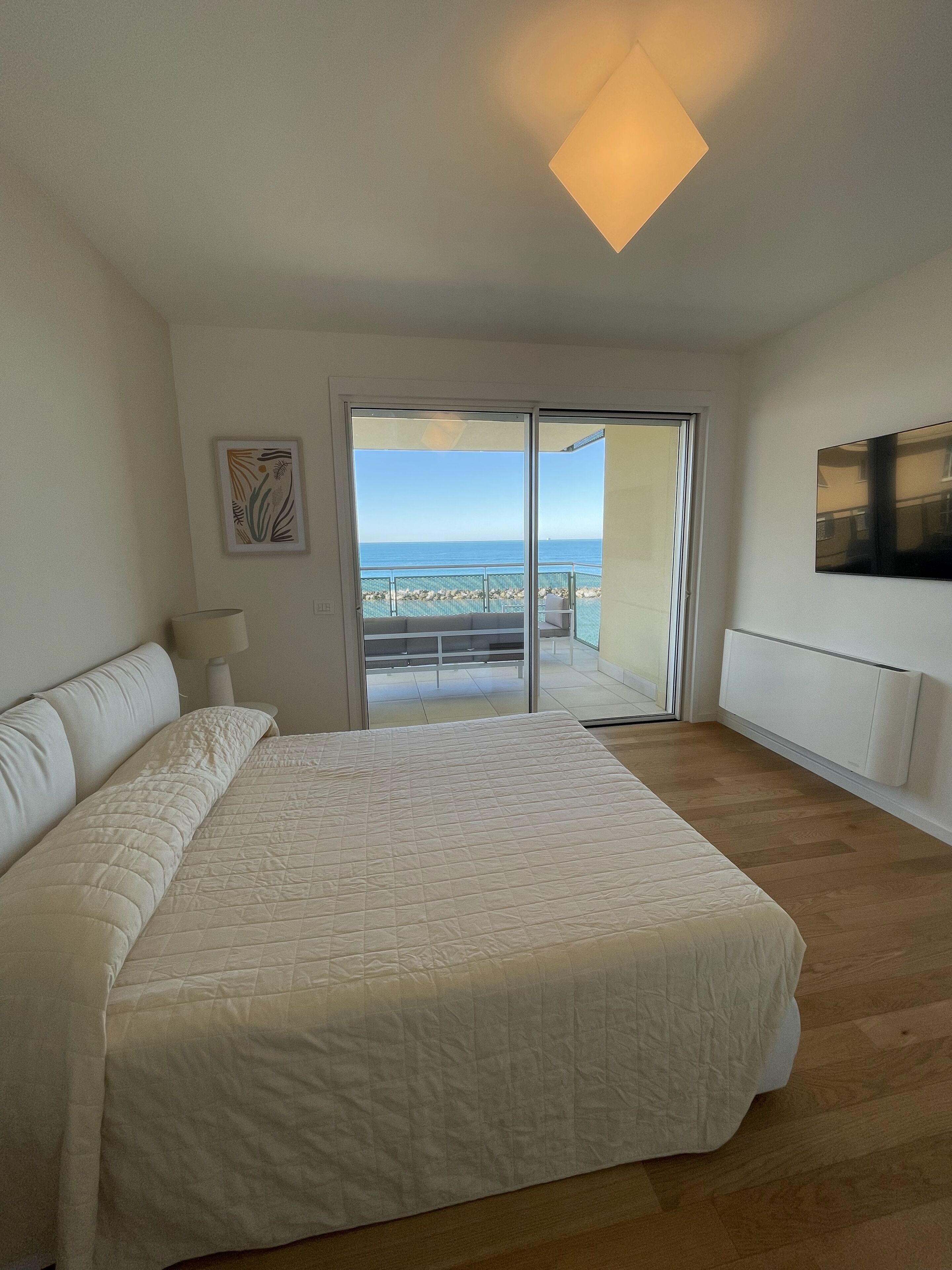 A bedroom with a large bed, a sliding glass door to a balcony, a mounted TV, and a ceiling light.