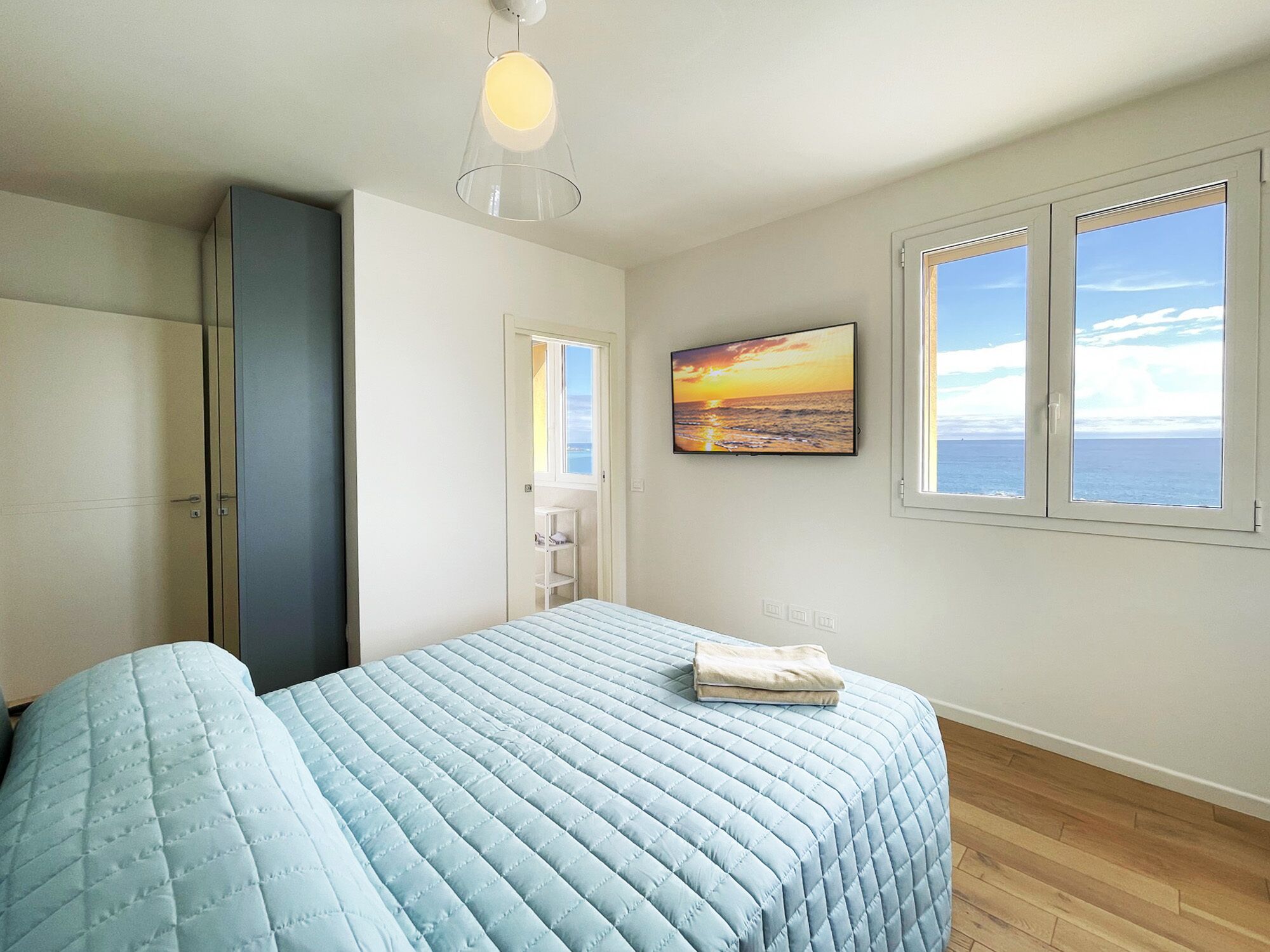 Um quarto com cama, guarda-roupa, pintura de um pôr do sol e janela vista para o mar.