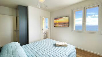Um quarto com cama, guarda-roupa, pintura de um pôr do sol e janela vista para o mar.