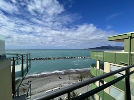 Vista costera desde un balcón con vista a una playa y un rompeolas rocoso, cielo despejado y montañas al lejano.