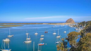 A coastal marina with numerous sailboats anchored in calm waters, a prominent rocky hill in the background, and a small settlement with buildings and greenery.
