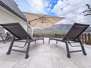 Une terrasse dotée de deux chaises longues et d’un grand parasol, offrant une vue sur un paysage montagneux.