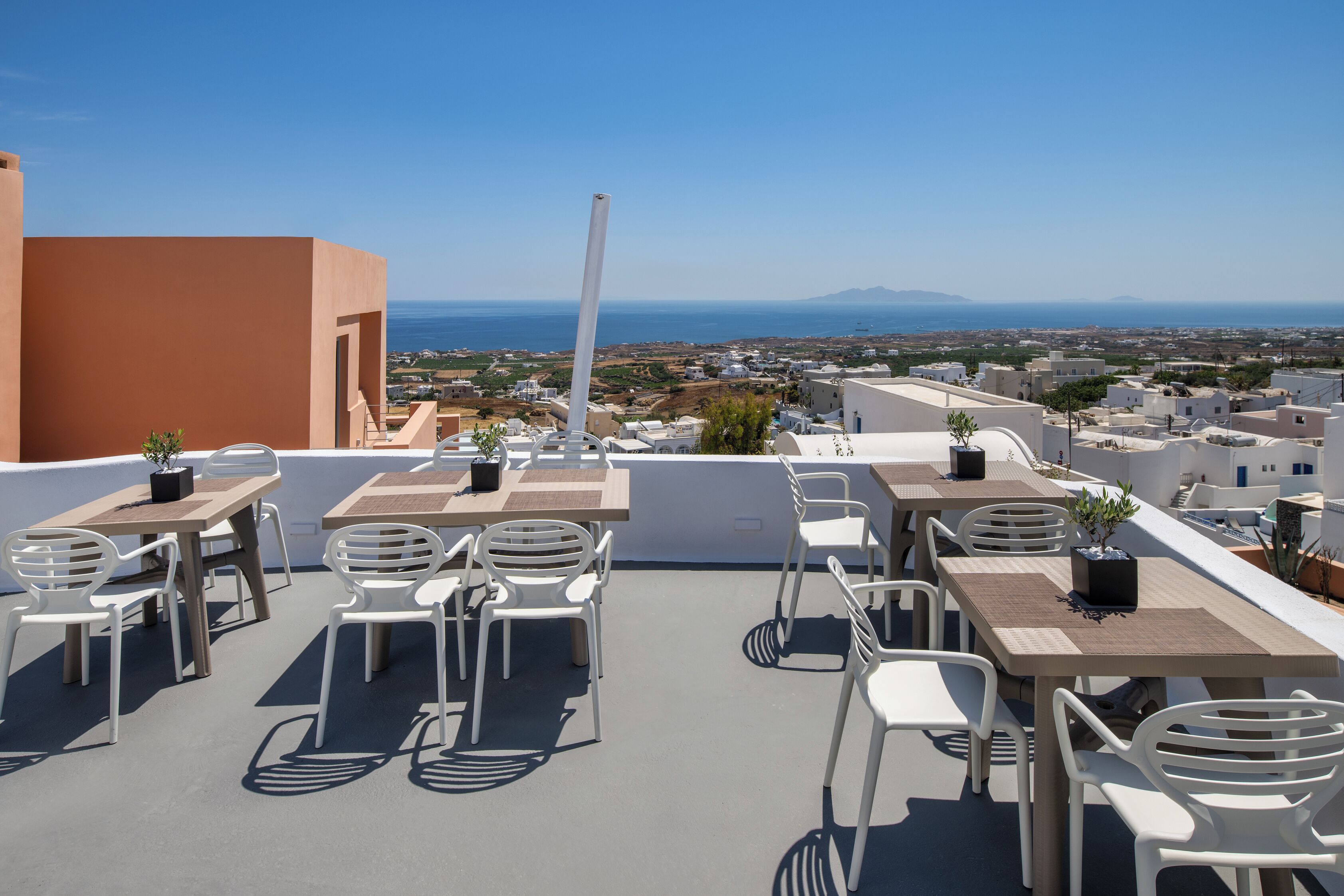 A rooftop terrace with white chairs and tables, overlooking a coastal cityscape with clear skies.