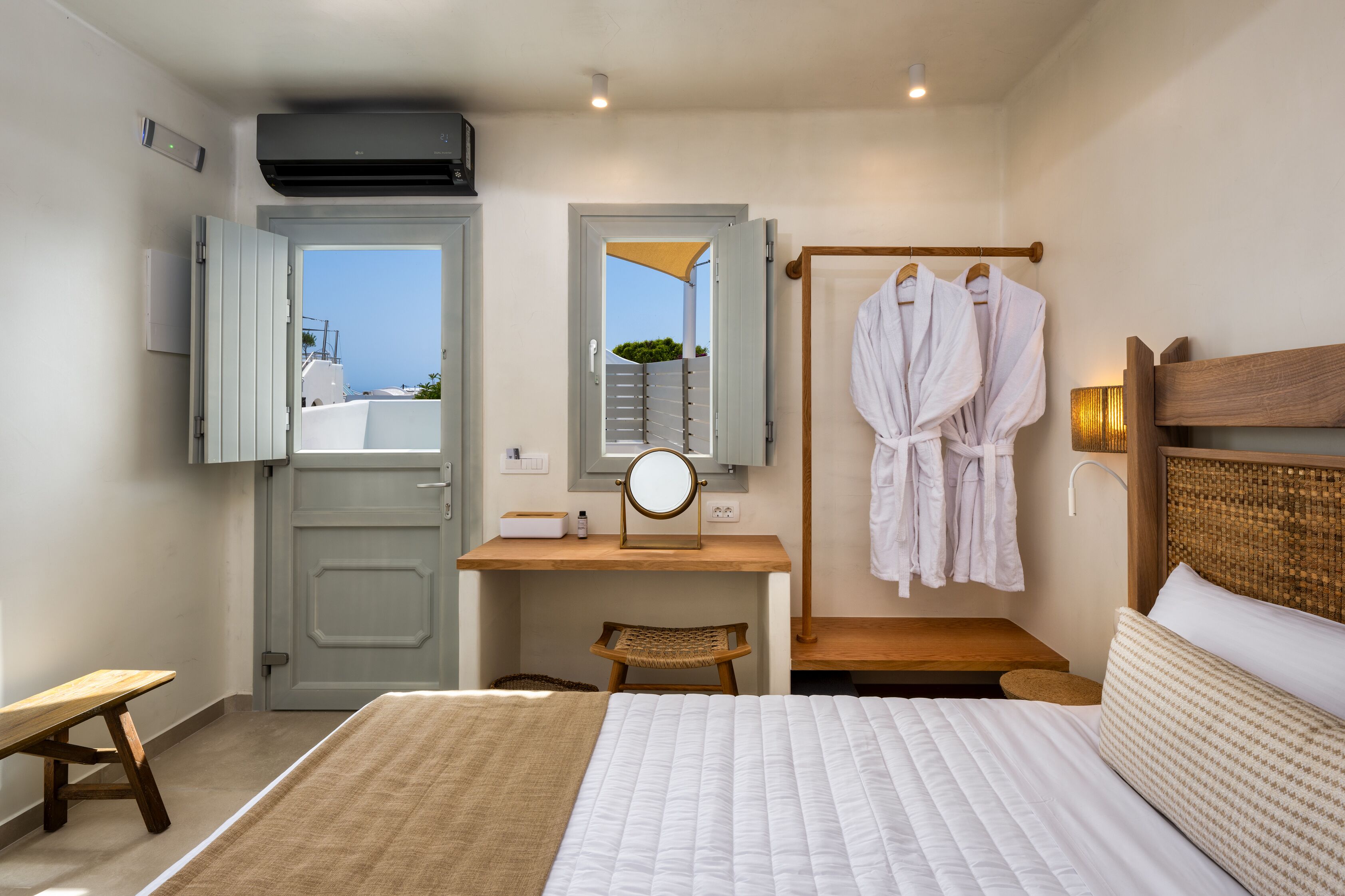A hotel room with a bed, a desk with a mirror, a chair, and a rack with robes.