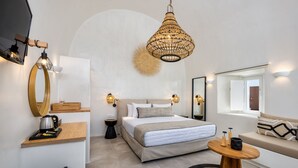 A modern bedroom with a large bed, bedside tables, a round wooden table, and a unique woven light fixture. - Hill Suites (Santorini)