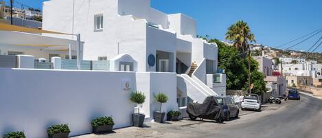 A white building with a staircase leading to a balcony, potted plants, and parked cars on a clear day.