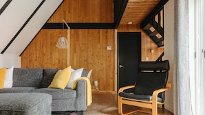 Un acogedor espacio tipo loft con techo de madera, un sofá seccional gris con cojines amarillos, un sillón de cuero negro y una puerta negra.