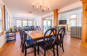 A spacious dining area with a large wooden table and black upholstered chairs, a chandelier, and a television in the background.