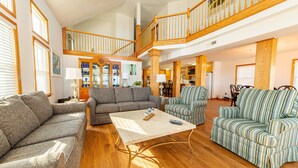 Ein geräumiger Wohnbereich mit hoher Decke, einer Holztreppe und verschiedenen Sitzgelegenheiten, darunter Sofas und Sessel.